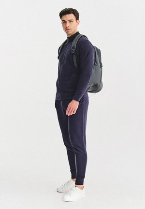 Mężczyzna ubrany w granatowy dres i białe trampki, noszący czarny plecak, stoi na jednolitym białym tle.