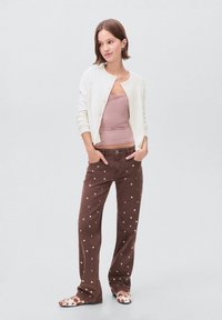 Tânără stând cu mâinile în buzunare, purtând un cardigan alb, un top roz, pantaloni maro cu capse argintii și pantofi cu imprimeu de vacă.