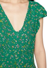 Grünes Blumenkleid mit tiefem V-Ausschnitt, kurzen, schwingenden Ärmeln und verstreuten rosa und gelben Blumenmustern auf einem strukturierten Stoff.