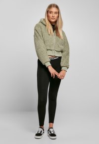 Geknipte olijfgroene nepbont hoodie met een voorrits; gecombineerd met zwarte legging. Model draagt zwarte sneakers met witte accenten.