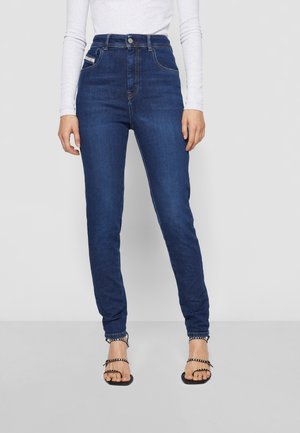 Femme portant un jean skinny taille haute bleu foncé et un haut blanc à manches longues, debout devant un fond clair uni.
