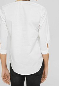 Chemise blanche à manches longues avec poignets relevables, texture lisse, ourlet arrondi et sans motifs visibles. Portée avec un pantalon noir ajusté.