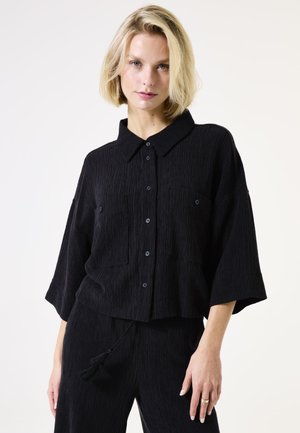 Camisa negra texturizada con cuello, botones en la parte delantera y dos bolsillos en el pecho. Presenta mangas anchas y un diseño acortado.