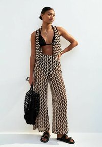 Un gilet court à motif zigzag noir et beige associé à un pantalon large, porté avec un haut de bikini noir et un sac en filet. Des sandales plates complètent le look.