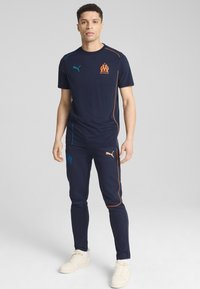 Navy blue Puma sportinė apranga, su oranžinėmis ir turkio akcentais. Marškiniai turi logotipą, o kelniuose logotipas yra ant klubo.