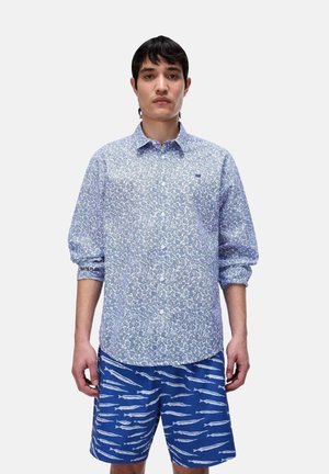 Junger Mann trägt ein blau-weißes langärmliges Hemd mit Blumenmuster und blaue Shorts mit weißen Fischmustern, steht vor einem einfarbigen Hintergrund.