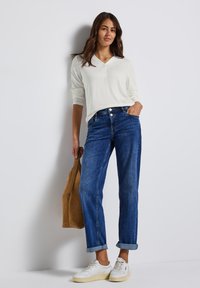 Weißes Langarmoberteil mit V-Ausschnitt, blaue Jeans mit aufgerolltem Saum und weiße Sneakers. Zubehör umfasst eine braune Tragetasche.