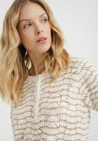Pull en tricot avec un motif rayé crème et beige, manches courtes et un boutonnage au niveau du col. Matière légère et texturée.