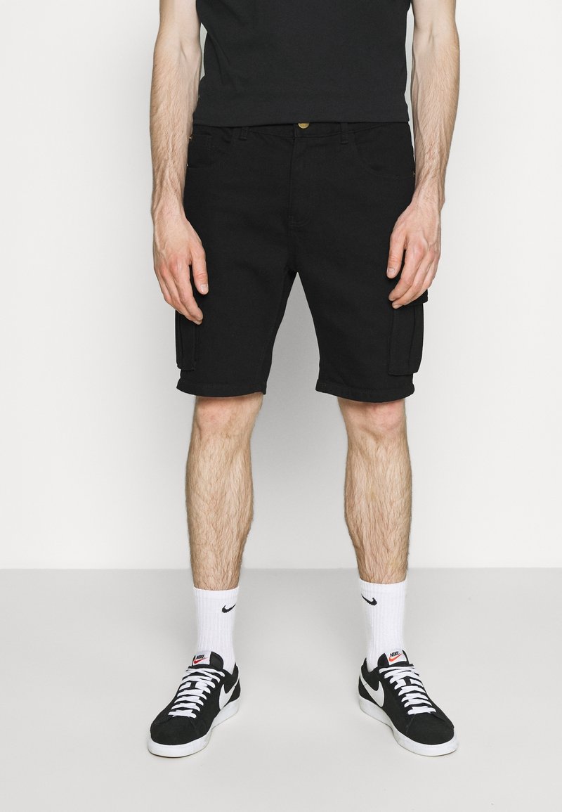 Černé cargo šortky s dvěma bočními kapsami, nastavitelným pasem a rovný střih. Nosené s černými teniskami a bílými ponožkami.