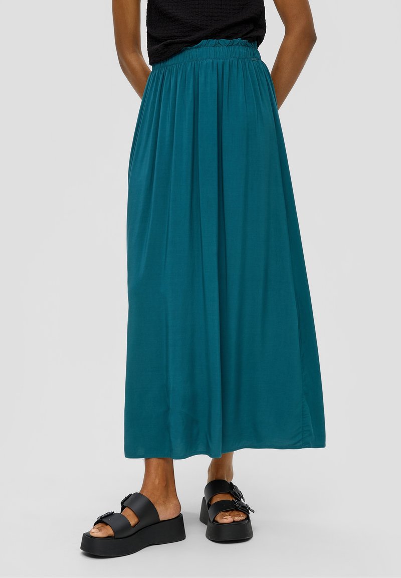 Jupe longue turquoise en tissu léger avec une taille élastique, présentant une texture lisse et un tombé fluide. Sandales à plateforme noires attachées.