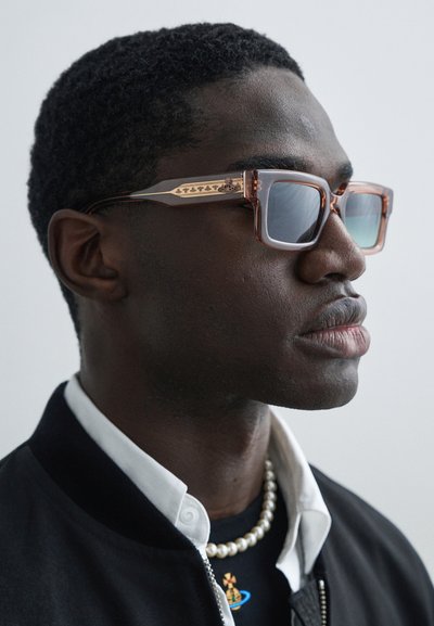 Τετράγωνα γυαλιά ηλίου με ελαφρώς καφέ σκελετό, φακούς με γκρι απόχρωση και χαραγμένες λεπτομέρειες στα κλασικά μέρη. Φορεμένα από άτομο με κοντό, στεγνό μαλλί.