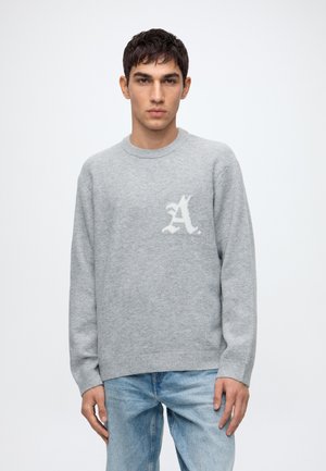 Jeune homme aux cheveux courts et foncés porte un pull gris clair avec la lettre blanche A sur la poitrine et un jean bleu clair, debout devant un fond blanc uni.