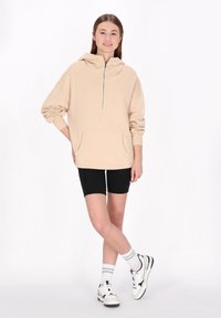Beige, oversized kapucnis pulóver fél cipzárral, kenguru zsebbel, bordázott mandzsettákkal, fekete bicikli sorttal és fekete akcentusokkal díszített fehér sportcipővel párosítva.