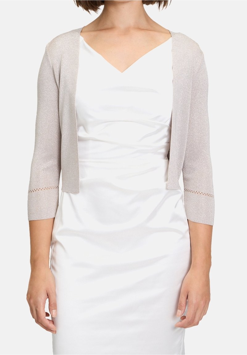Ljusgrå metallic stickad bolerojacka med trekvartslånga ärmar och spetsliknande kant vid ärmslutet, bärs över en vit satinklänning.