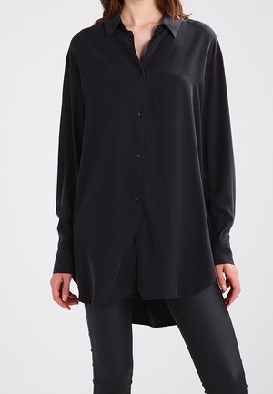 Kvinde iført en sort skjorte med lange ærmer og knapper kombineret med tætsiddende sorte bukser, stående foran en ensartet lys baggrund.