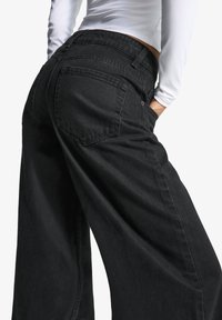 Črne široke jeans hlače iz denima, z zadnjimi žepi, visokim pasom in gladko teksturo. Podrobnosti vključujejo zanke za pas in šive.