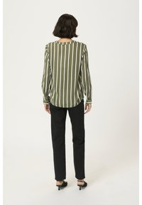 Femme de dos portant un chemisier rayé blanc et olive, un pantalon noir et des chaussures à talons noires sur un fond blanc.