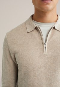 Bezs kötött pulóver gallérral és negyed cipzárral. Texturált felülettel és réteges stílusban világos színű pólóval alatta.