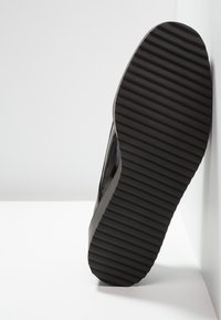 Semelle de chaussure avec des rainures horizontales parallèles, en caoutchouc noir, inclinée contre une surface blanche.