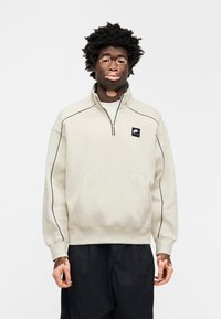 Μπεζ φούτερ με φερμουάρ μέχρι το μέσο του στήθους και μαύρες λεπτομέρειες περιμετρικά, με μπροστινή τσέπη και ένα μικρό μαύρο λογότυπο. Φορεμένο με σκουρόχρωμο παντελόνι.