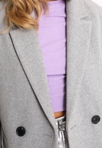 Grå uldblazer med sorte knapper og indsatsklapper. Underneath ses en lilla ribbet top, der fremhæver det lagdelte look.