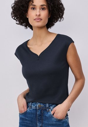 Femme aux cheveux bouclés portant un haut sans manches bleu marine foncé et un jean bleu, les mains dans les poches, sur un fond uni.