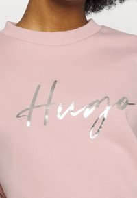 Rosa Sweatshirt mit Rundhalsausschnitt und einem silbernen, metallischen "Hugo"-Logo auf der Vorderseite. Das Material wirkt glatt mit einer sanften Textur.