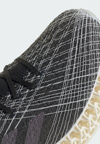 Black athletic shoe with a textured knit upper featuring white diagonal lines, black laces, and a distinctive cream-colored sole design.