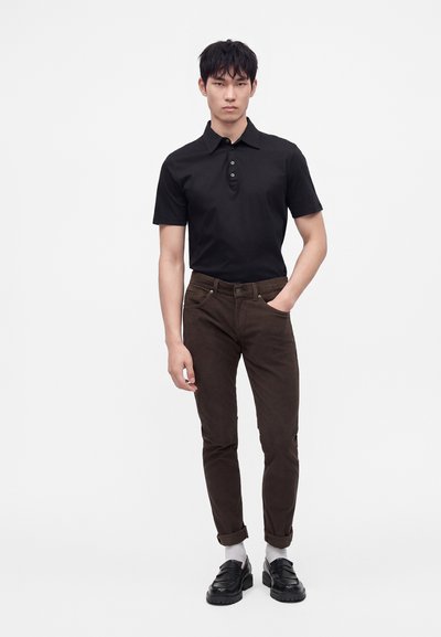 Jeune homme debout, une main dans la poche, portant un polo noir à manches courtes, un pantalon marron foncé retroussé aux chevilles, des chaussettes blanches et des chaussures noires.