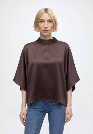 Femme aux cheveux blonds courts portant un haut en satin marron foncé ample et un jean bleu, debout devant un fond clair uni.