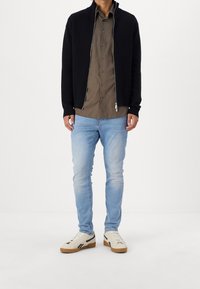 Svart zip-up tröja med ribbade muddarna, brun skjorta med knappar, ljusblå jeans och beige sneakers med svarta accenter. Enkel, avslappnad klädsel.