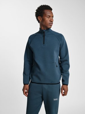 Man in donkerblauwe sweatshirt met kwart rits en bijpassende broek, staand en naar de zijkant kijkend tegen een effen lichte achtergrond.