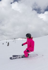 Skiausrüstung in leuchtendem Pink, bestehend aus einer Jacke und einer Hose. Die Skier sind dunkel mit Bindungen. Verschneite Piste und bewölkter Himmel im Hintergrund.