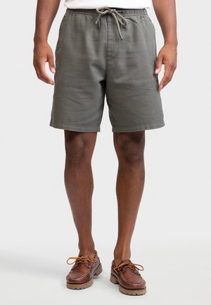 Mies, jolla on harmaat kiristysnauhalliset shortsit ja ruskeat nahkaiset purjehduskengät, seisoo yksinkertaisen taustan edessä.