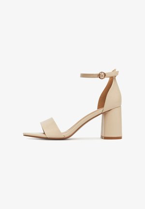 Beżowy sandał na wysokim obcasie z kwadratowym noskiem, szerokim paskiem z przodu oraz regulowanym paskiem wokół kostki, z grubym obcasem i gładką teksturą.