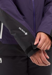 Veste imperméable violette avec manches noires, dotée d'un poignet à velcro et du logo "Gore-Tex". Fermeture éclair à l'avant et tissu corporel texturé.