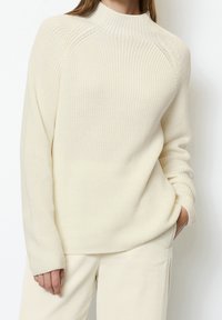 Pull en maille couleur crème avec col montant, texture côtelée et manches raglan. Associé à un pantalon côtelé de couleur claire assorti.