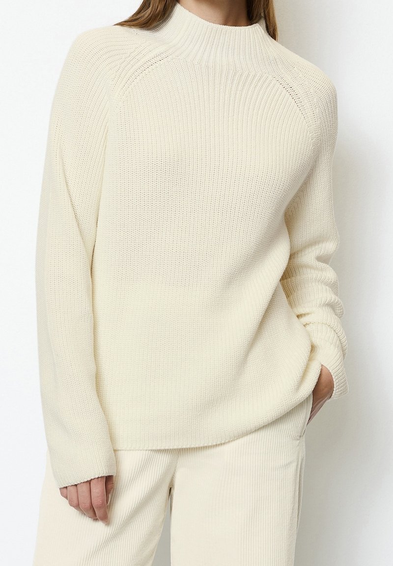 Pull en maille couleur crème avec col montant, texture côtelée et manches raglan. Associé à un pantalon côtelé de couleur claire assorti.