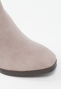 Gros plan sur une botte cheville en daim beige avec un bout arrondi et une semelle marron foncé sur un fond blanc.