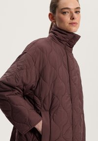 Gequilted jas in diep bordeaux, met een hoge kraag, oversized ontwerp en een soepele textuur met zichtbare gebogen stikdetails.