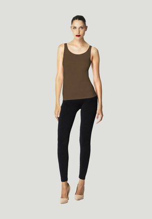 Donna in piedi che indossa una canotta marrone, leggings neri e tacchi beige su uno sfondo grigio chiaro.