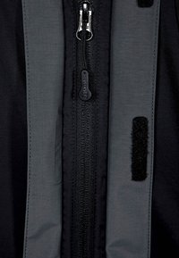 Černá bunda se středním zipem, vyznačující se šedými látkovými akcenty a suchým zipem. Hladký povrch a robustní konstrukční detaily.