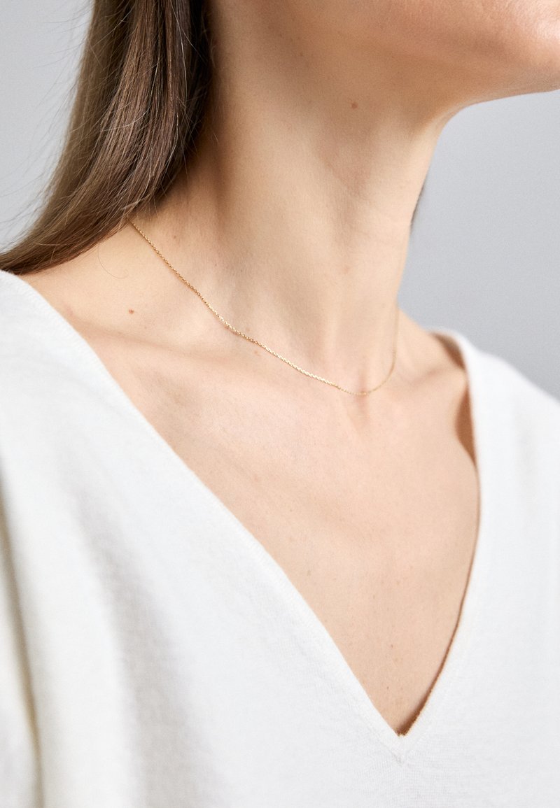Culoare de aur, lănțișor cu zale fine, purtat pe un gât gol, asortat cu o piesă vestimentară albă, cu decolteu în V. Textură netedă, design minimalist.