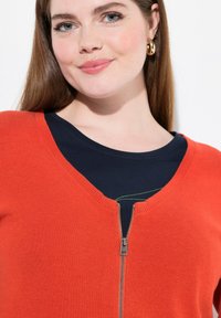 Nainen, joka on pukeutunut tummansiniselle paidalle oranssin vetoketjupuseron alle, pitkät ruskeat hiukset ja kultaiset renkaakorvakorut, hymyilee lempeästi.