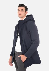 Giacca impermeabile blu navy con cappuccio, chiusura frontale con zip, tasche laterali e texture del tessuto liscia, indossata sopra una camicia bianca e pantaloni grigi.