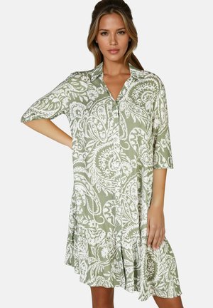 Femme portant une robe chemise boutonnée à motifs cachemire verts et blancs avec col et manches trois-quarts, posant avec une main sur la hanche.