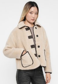 Beige fleece jacket with black trim, featuring a collared neckline, five button closures, and two front pockets. Soft, textured fabric.
