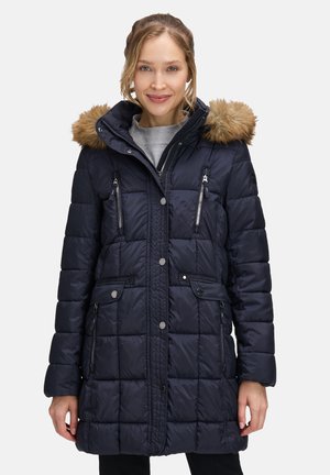 Kobieta w granatowym pikowanym płaszczu zimowym z kapturem wykończonym futrem, z zamkiem błyskawicznym i zapięciem na napy z przodu oraz z wieloma kieszeniami na zamek.