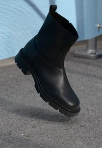 Black leather ankle boot with rugged sole and pull tab, suspended above asphalt ground with a blue textured background.