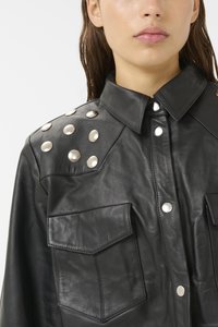 Zwart leren overhemdjasje met een kraag, voorzien van zilveren drukknopen en zes metalen studs op de schouders. Twee zichtbare borstzakken.